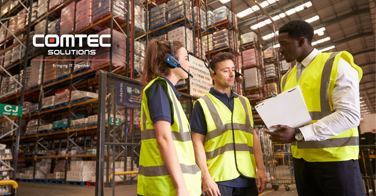 Three workers in vests and headsets stand in a warehouse. One holds a clipboard. 'COMTEC SOLUTIONS – Bringing IT Together' appears above.